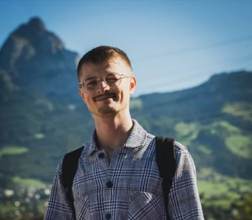 Elliot zwinkert in die Kamera, im karierten Hemd vor Bergen