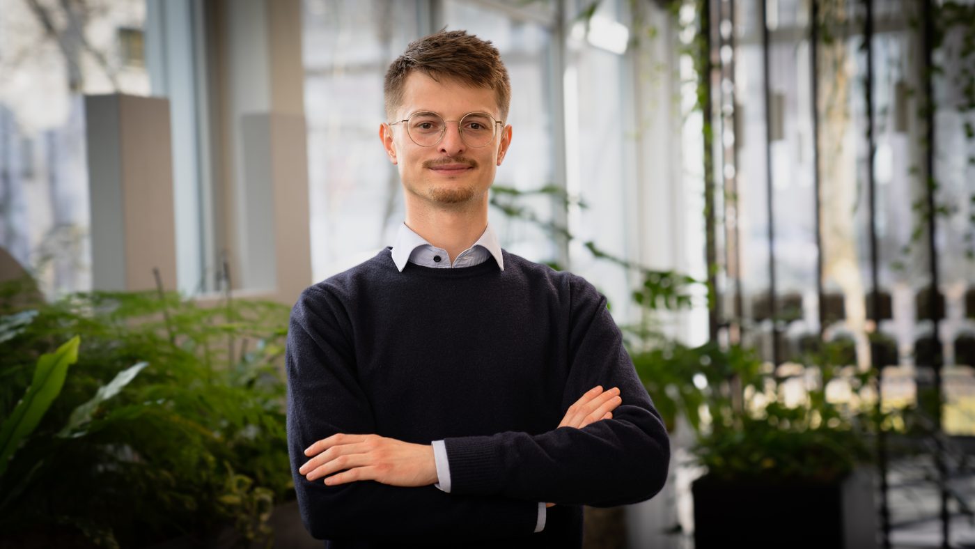 Elliot Schneider, Porträt mit verschränkten Armen vor einer Fensterfront mit Pflanzen